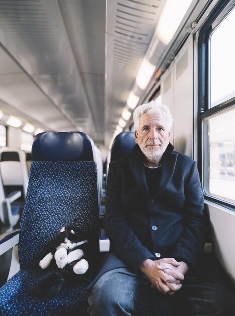 Ein älterer Mann sitzt neben einer schwarzen und weißen Katze im Zug. | © Ingo Pertramer