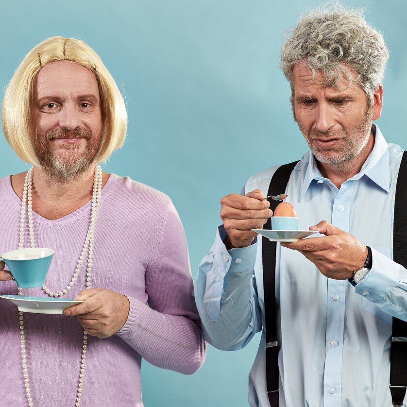 Zwei Männer, einer mit blonder Perücke und der andere nachdenklich, genießen eine Tasse Tee und ein weich gekochtes Ei. | © Udo Leitner