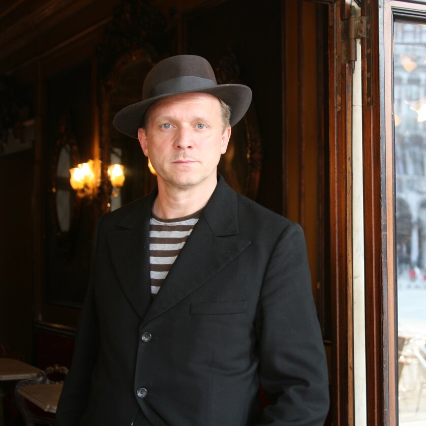 Ein Mann in schwarzem Outfit und Hut steht vor einem offenen Fenster. | © Katharina John