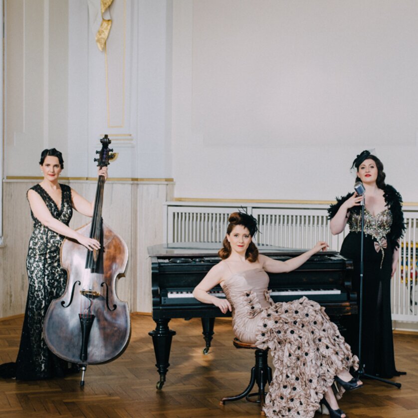 Drei Musikerinnen posieren in eleganten Kleidern vor einem Klavier. | © ladiesandlord