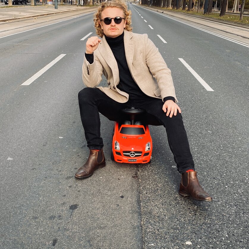 Ein Mann sitzt auf einem roten Spielzeugauto in einer leeren Straße. | © MutterBohl