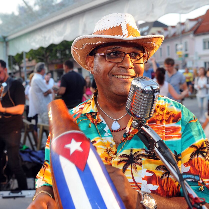 Latin Live am Lendplatz | © Latin Live Ein Musiker in bunter Hawaii-Hemd und Strohhut spielt bei einem Straßenfest vor einem Mikrofon. Er hält ein Instrument mit der kubanischen Flagge, im Hintergrund spielen weitere Musiker und Menschen tanzen. Die Stimmung ist fröhlich und lebendig. | © Latin Live