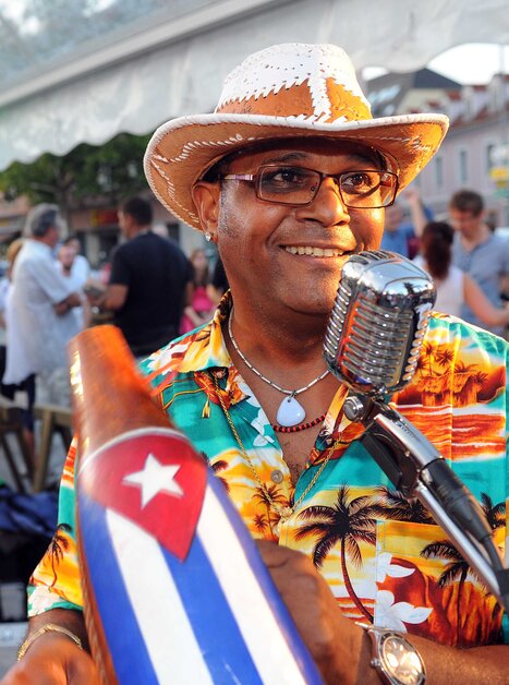 Ein Musiker in bunter Hawaii-Hemd und Strohhut spielt bei einem Straßenfest vor einem Mikrofon. Er hält ein Instrument mit der kubanischen Flagge, im Hintergrund spielen weitere Musiker und Menschen tanzen. Die Stimmung ist fröhlich und lebendig. | © Latin Live