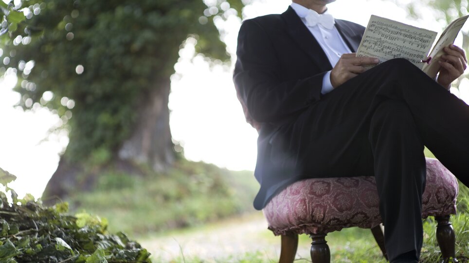 Ein eleganter Mann im Anzug liest Notenblätter auf einem gepolsterten Stuhl. | © Pilo Pichler