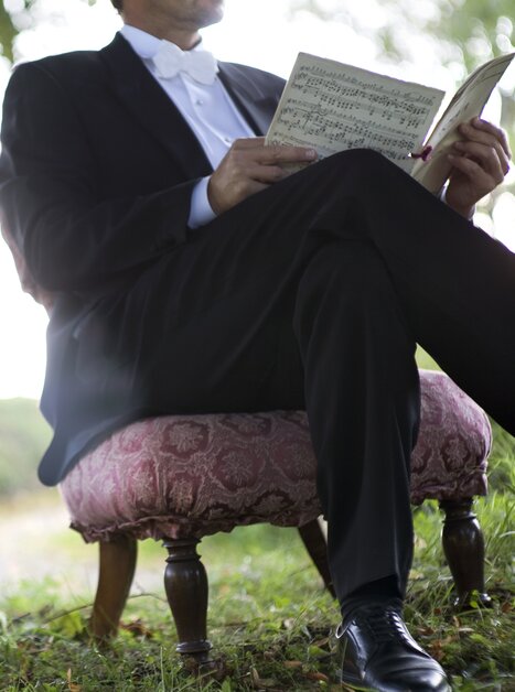 Ein eleganter Mann im Anzug liest Notenblätter auf einem gepolsterten Stuhl. | © Pilo Pichler