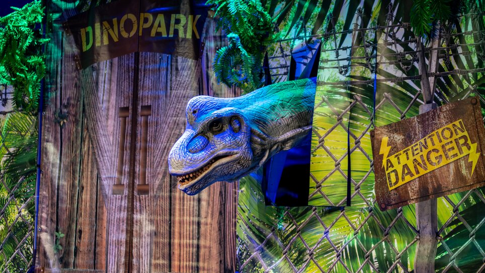 Dinosaurierkopf schaut aus einer Kulisse bei einer Dinosaurier-Show. | © Timo Tutschek