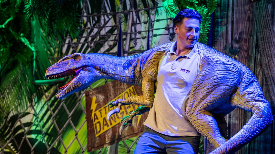 Ein junger Mann posiert mit einer Dinosaurier-Puppe auf einer Bühne bei einer Dinosaurier-Show. | © Timo Tutschek