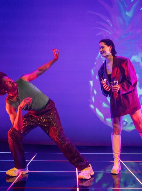 Zwei Darsteller interagieren auf einer Bühne mit farbenfrohen Lichtern. | © Phile Deprez