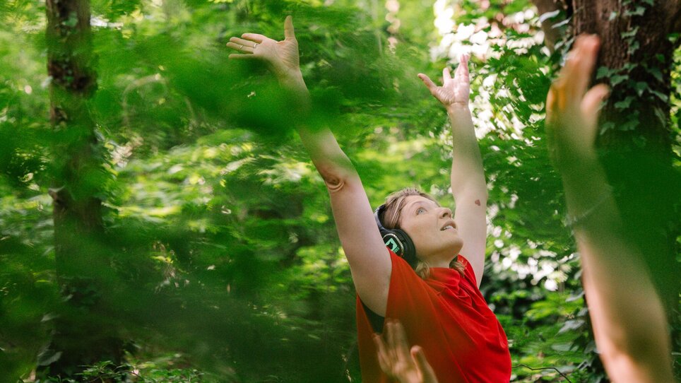 Frau hebt die Arme im Wald, hört Musik mit Kopfhörern. | © Alek Kawka