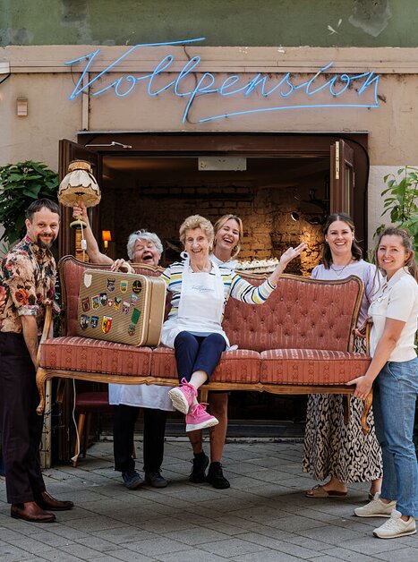 Sechs fröhliche Menschen vor dem Café „Vollpension“, fünf heben ein Sofa mit einer älteren Frau darauf. Alle lachen, die Szene wirkt lebendig und herzlich. | © Oma on Tour
