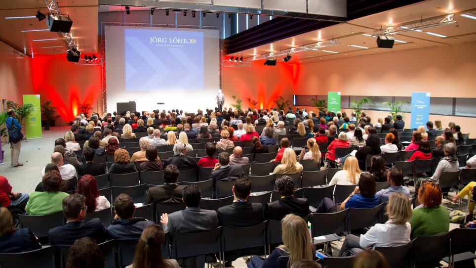 Large gathering in a modern conference hall with attendees. Jörg Löhr speaking on stage. | © MCG - Martin Wiesner