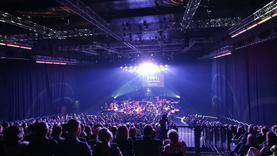 Orchester spielt in einem abgedunkelten Saal mit Publikum. | © Helmut List Halle