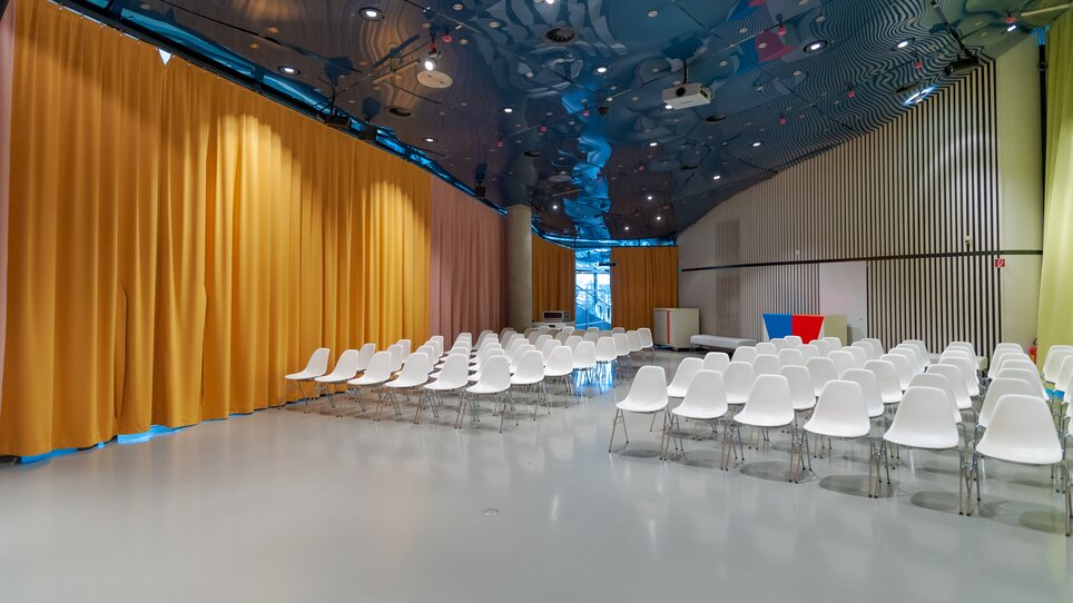 Spacious room with a high, reflective ceiling in blue tones, white chairs arranged in rows facing forward, and a large golden curtain on one side; clean, modern atmosphere with flexible event setup. | © UMJ -N. Lackner