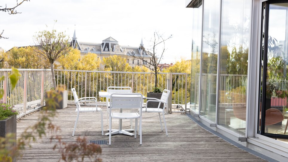 Eine moderne Dachterrasse mit weißen Stühlen und Tisch bietet eine entspannte Atmosphäre im Level 4. Hinter der Dachterrasse sieht man die bunten Herbstbäume und die Architektur von Graz. | © Level 4