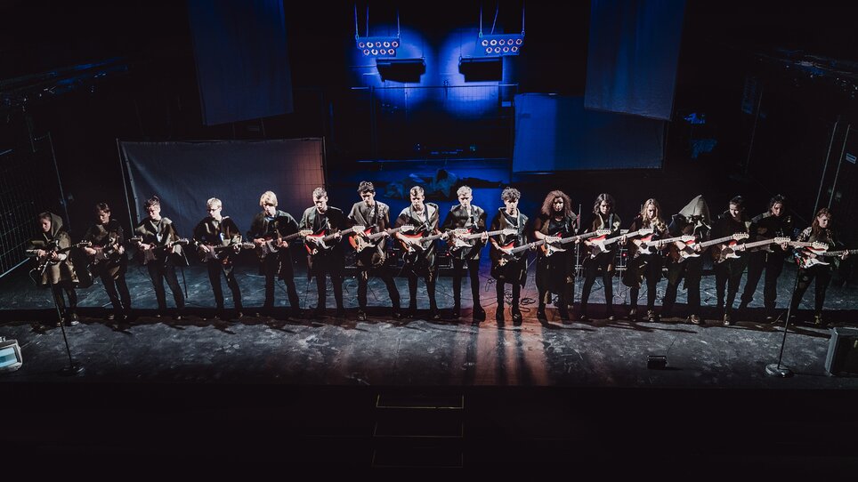 Eine Gruppe von Musikern im 'Next Liberty' spielt auf E-Gitarren. Die Bühne ist modern beleuchtet, was eine kreative Atmosphäre schafft. | © Lupi Spuma