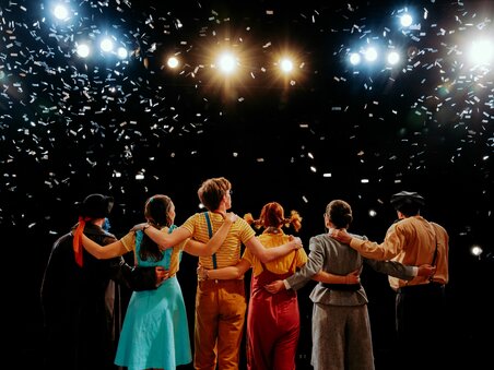 Die Bühne von Next Liberty zeigt eine Gruppe von Darstellern während der Schlussapplaus. Es fallen Konfetti und das Licht der Scheinwerfer erzeugt eine festliche Atmosphäre. | © Stella Kager