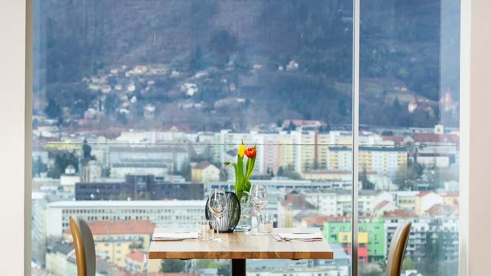 In Restaurant SCHLOSSBERG in Graz, a stylish table and two chairs offer a view of the city. Large windows provide an inspiring outlook on the Mur and surroundings. Modern design and ample lighting create a welcoming atmosphere for business trips and events. | © Restaurant SCHLOSSBERG - Paul Stajan