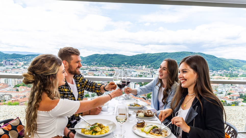Nel Restaurant SCHLOSSBERG, quattro persone sono sedute attorno a un tavolo con vista su Graz. Brindano con bicchieri di vino e godono di un pasto elegante in un ambiente moderno. | © Restaurant SCHLOSSBERG - Paul Stajan