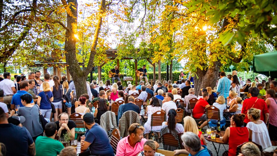 Das Restaurant SCHLOSSBERG zeigt eine lebhafte Atmosphäre mit vielen Gästen. Gitarrenspieler treten auf einer Außenterrasse unter Bäumen auf. | © Restaurant SCHLOSSBERG - Werner Krug