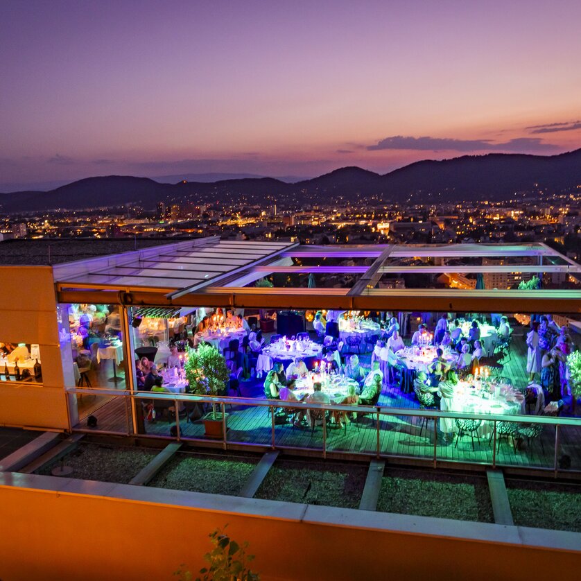 Auf der Dachterrasse des Restaurant SCHLOSSBERG genießen Gäste ein festliches Dinner mit Blick auf Graz bei Dämmerung. Die eleganten Tische sind stilvoll gedeckt und von Pflanzen umgeben, während die moderne Architektur und die bunten Lichter eine einladende Atmosphäre schaffen. | © Restaurant SCHLOSSBERG - Werner Krug