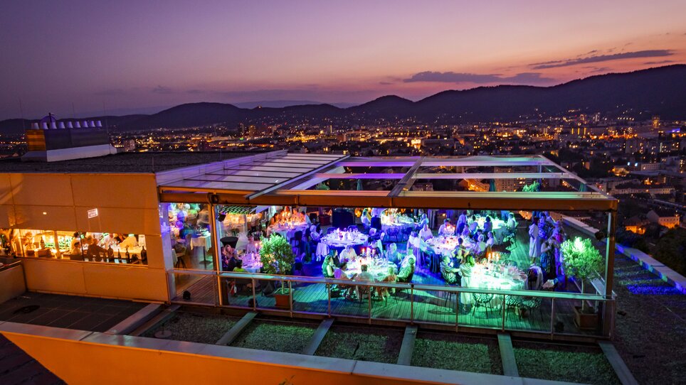 Auf der Dachterrasse des Restaurant SCHLOSSBERG genießen Gäste ein festliches Dinner mit Blick auf Graz bei Dämmerung. Die eleganten Tische sind stilvoll gedeckt und von Pflanzen umgeben, während die moderne Architektur und die bunten Lichter eine einladende Atmosphäre schaffen. | © Restaurant SCHLOSSBERG - Werner Krug