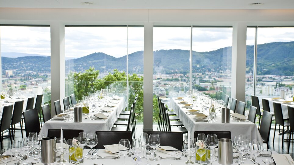 Im Restaurant SCHLOSSBERG werden stilvolle Tischanordnungen mit Blick auf die Stadt Graz und die Mur präsentiert. Die moderne Architektur und großzügigen Fenster schaffen eine helle, einladende Atmosphäre für Konferenzen und Business-Events. | © Restaurant SCHLOSSBERG