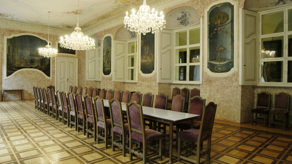 Ein eleganter Raum in Stift Rein mit langen Tafeln, Kronleuchtern und künstlerischer Wandgestaltung, ideal für Tagungen und Konferenzen.