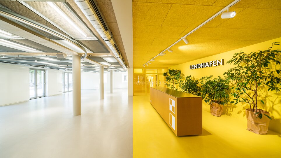 Der Lendhafen in Graz bietet helle, moderne Räume mit beeindruckender Deckenhöhe und flexibler Möblierung. Die gelbe Farbgestaltung und Pflanzen schaffen eine einladende Atmosphäre für Konferenzen und Geschäftsveranstaltungen. | © Lupi Spuma