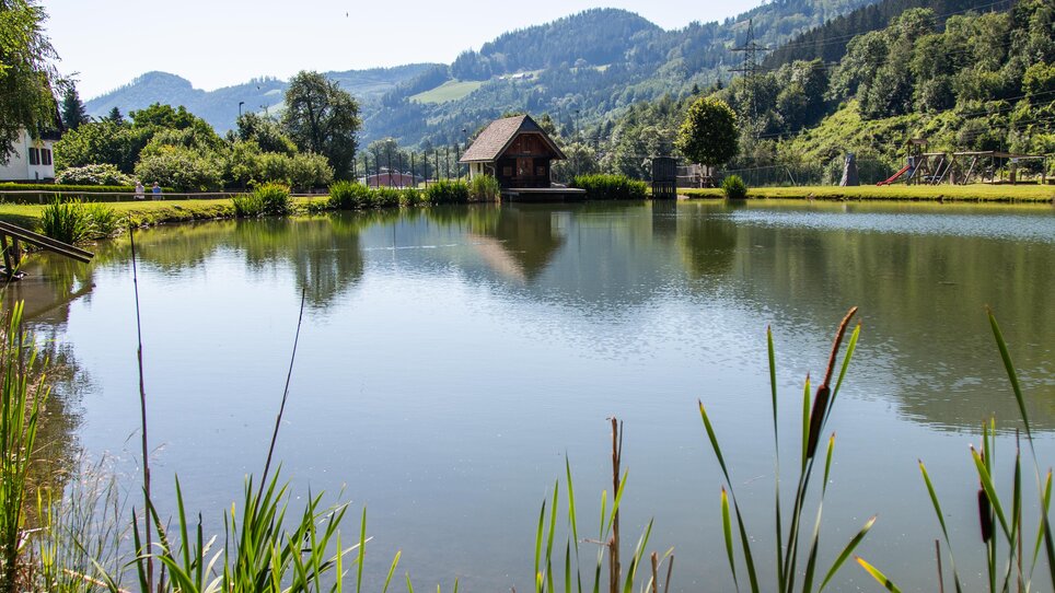 Ein ruhiger See umgeben von Bäumen und Hütten bei strahlendem Wetter. | © Grossauer