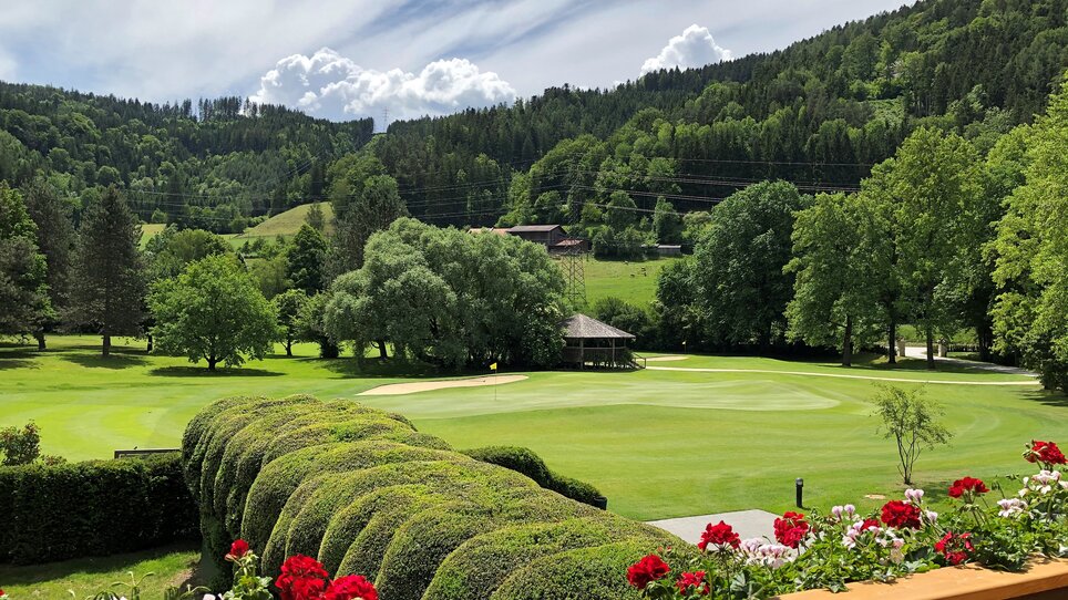 Grüner Golfplatz umgeben von Bäumen und Blumen, im Hintergrund Berge und Wolken beim Murhof Hotel & Restaurant. | © Murhof