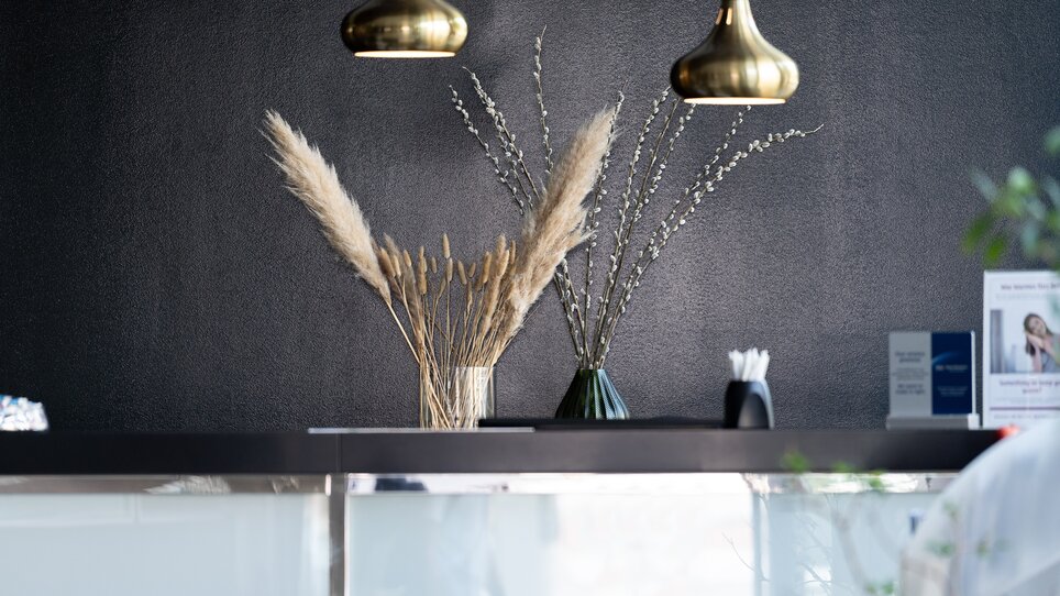 Empfangstheke mit Trockenblumen und goldenen Lampen im PLAZA INN Graz City. | © PLAZA INN Graz City