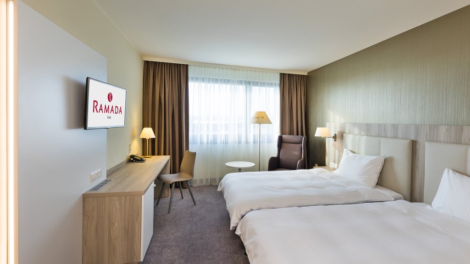 Hell erleuchtetes Hotelzimmer mit zwei Betten, Tisch und Sessel im Hotel Ramada Graz. | © Hotel Ramada Graz