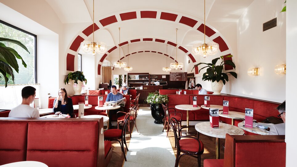 Interior view of a café in Hotel Das Weitzer with red upholstery, tables, and guests chatting. | © Florian Weitzer Hotels & Restaurants