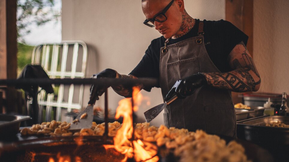 A chef of The Underground Kitchen // crew works focused at the grill with flames and prepared dishes in The Underground Kitchen //. | © Alex Rauch