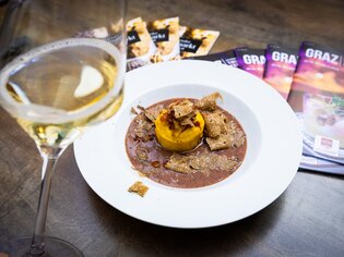 Trüffelfestival Graz - ein Trüffelgericht auf einem weißen Teller, ein Glas Weißwein und Prospekte vom Trüffelfestival auf einem braunen Tisch. | © Graz Tourismus - Werner Krug