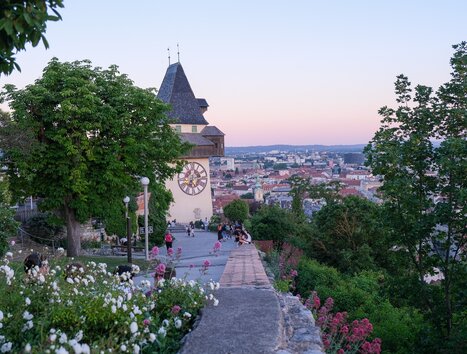 The Graz clock tower overlooking the city of Graz at sunset, surrounded by trees and flowers. | © putopis - Jasminka Dagovic