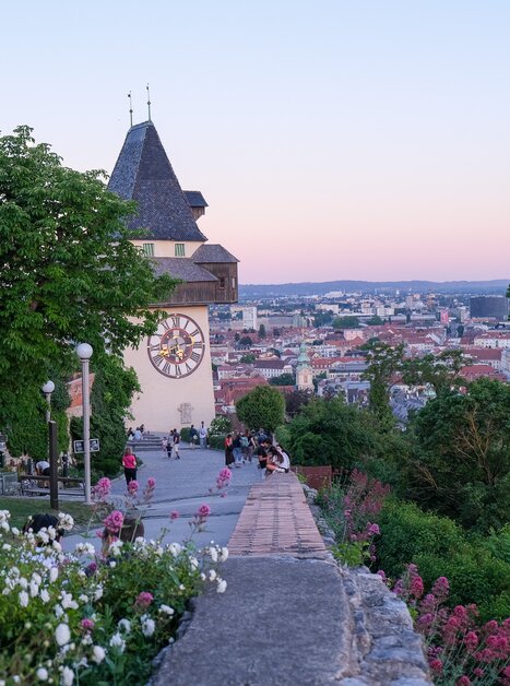 Der Grazer Uhrturm über der Stadt Graz bei Sonnenuntergang, umgeben von Bäumen und Blumen. | © putopis - Jasminka Dagovic