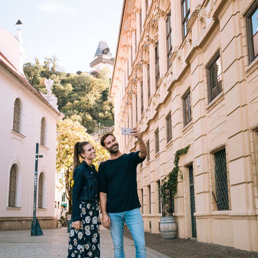 A couple is taking a selfie in the city of Graz, with the Graz Clock Tower in the background. | © Graz Tourismus