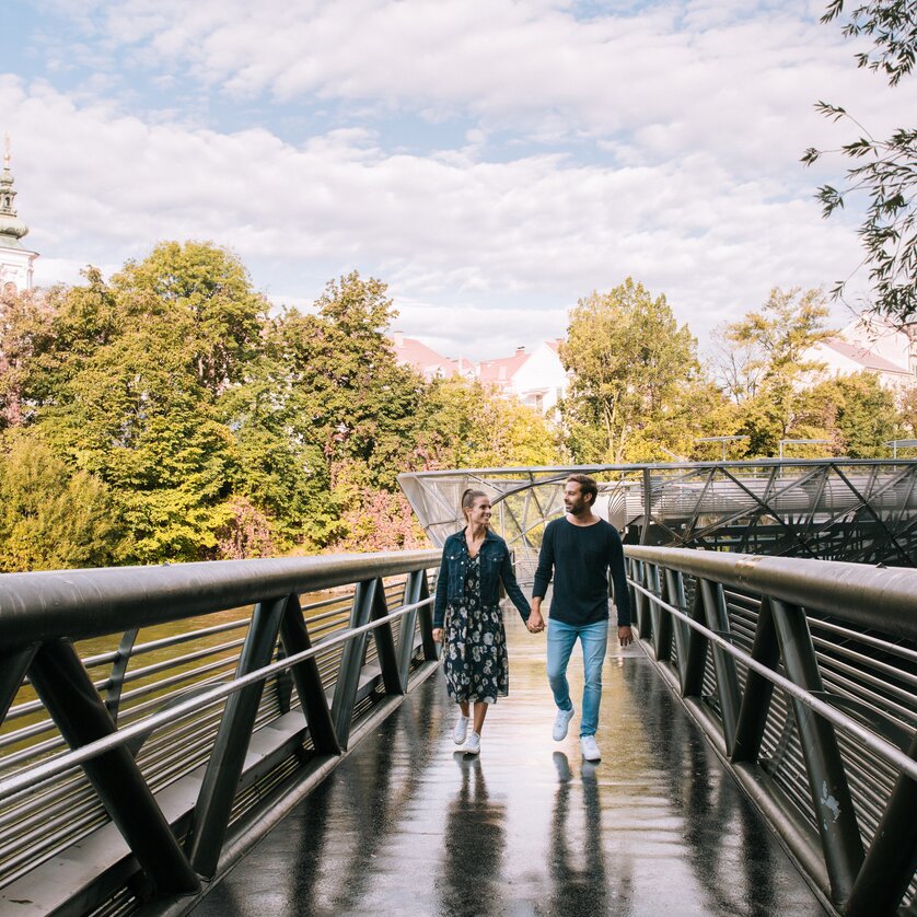 Paar spaziert auf der Murinsel in Graz, umgeben von Bäumen. | © Graz Tourismus