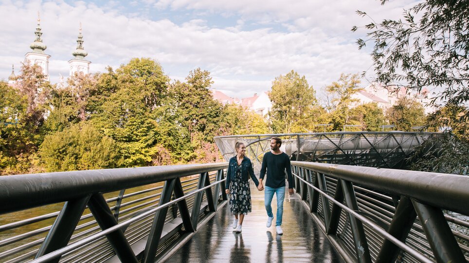 Coppia che cammina sull'Isola sul fiume Mur a Graz, circondata da alberi. | © Graz Tourismus