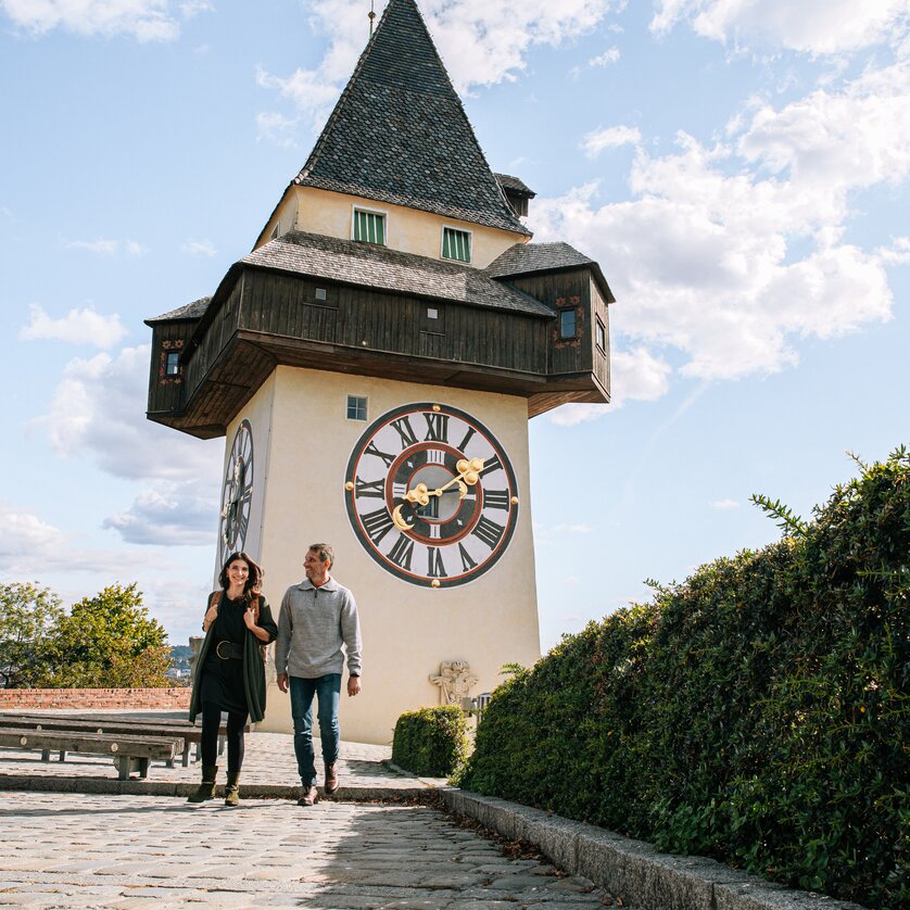 Couple walking in front of the Graz clock tower under a blue sky. | © Graz Tourismus