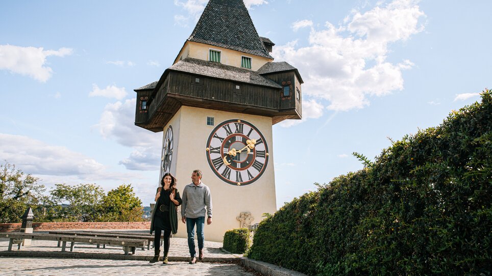 Coppia a passeggio davanti alla torre dell'orologio di Graz sotto un cielo azzurro. | © Graz Tourismus