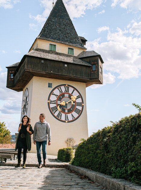Paar spaziert vor dem Grazer Uhrturm bei blauem Himmel. | © Graz Tourismus