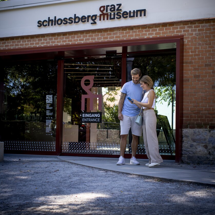 Paar steht am Eingang des Graz Museums Schlossberg und schaut auf das Handy. | © Graz Tourismus - Tom Lamm