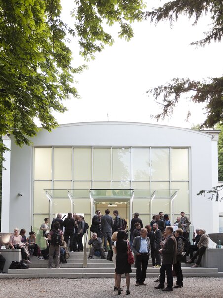 People gather in front of a modern white building with large windows. | © Maria Schnabl