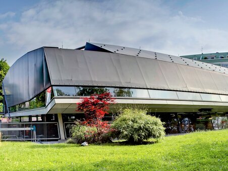 The image features a modern building in Graz, surrounded by green grass and trees. | © Hannes Loske