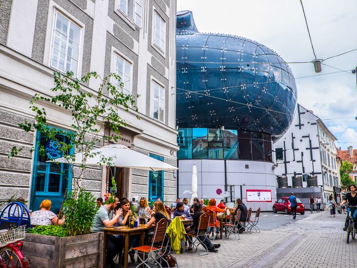 Blick auf das Kunsthaus Graz sowie ein Straßencafé mit Gästen. | © Becki Enright_Boarders of Adventure