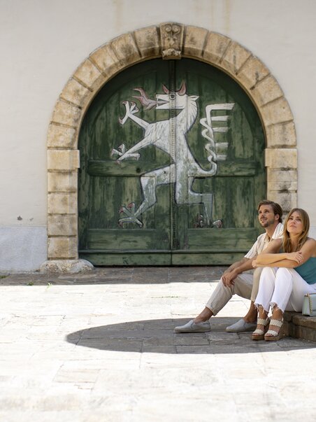 Couple sitting relaxed in Landhaus Courtyard in Graz in front of a colorful door. | © Graz Tourismus - Tom Lamm