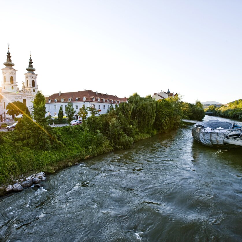 View of the Mur island in Graz, surrounded by trees and historic buildings.