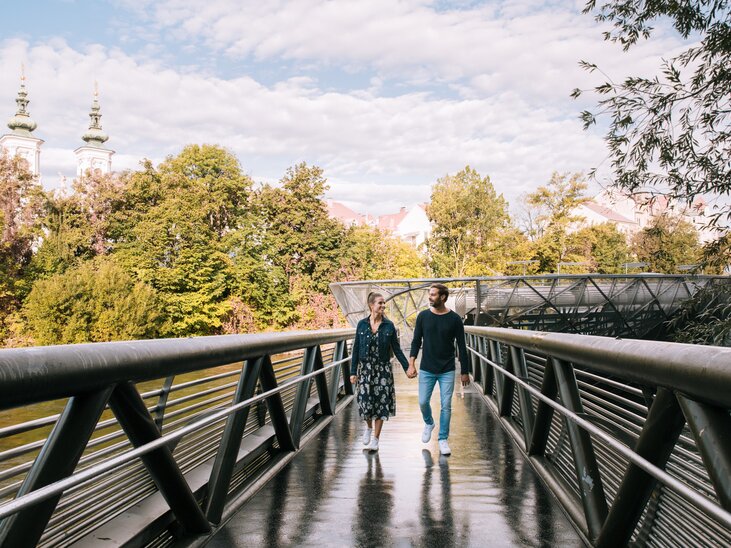 Paar spaziert auf der Murinsel in Graz, umgeben von Bäumen. | © Graz Tourismus